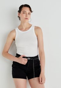 Bijela rebrasta majica bez rukava, crne kratke hlače viskog struka, srebrni pojas s dekorativnim detaljima. Model ima kratku, valovitu kosu i naušnice na obruče.