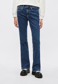 Vaqueros acampanados azul denim con lavado medio, cinco bolsillos y costuras visibles, combinados con zapatos negros de suela gruesa.