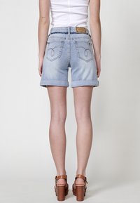 Shorts en denim bleu clair avec ourlets retroussés, présentant deux poches arrière et des coutures décoratives. Associés à des sandales à plateforme marron.