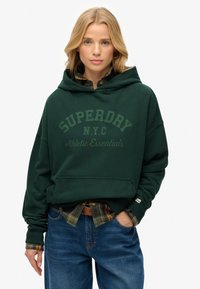 Sudadera verde oscura con un gran logo de "SUPERDRY", capucha ajustable, bolsillo frontal y puños acanalados, llevada sobre una camisa de cuadros y jeans de mezclilla.