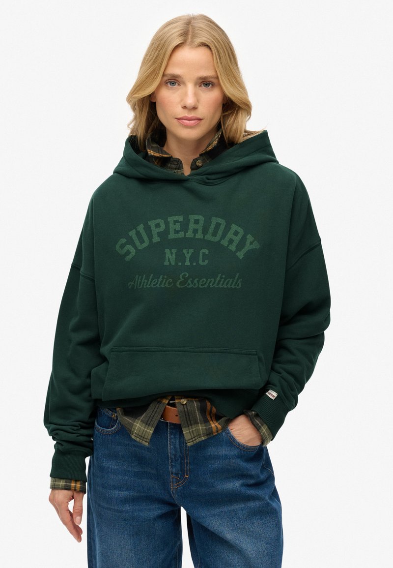 Sudadera verde oscura con un gran logo de "SUPERDRY", capucha ajustable, bolsillo frontal y puños acanalados, llevada sobre una camisa de cuadros y jeans de mezclilla.