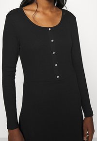 Černý bodysuit s dlouhým rukávem a kulatým výstřihem, s řadou pěti dekorativních knoflíků vpředu. Hladký textilní vzor.