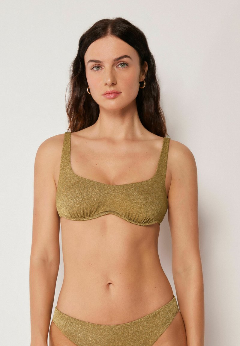 Femme aux longs cheveux foncés portant un haut et un bas de bikini pailletés dorés, debout devant un fond clair uni.