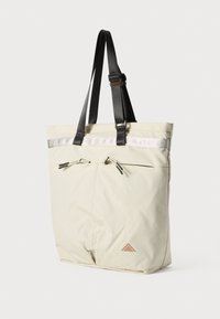 Sac fourre-tout en tissu crème, doté de poignées noires, d'une bande réfléchissante argentée, de deux zips sur le devant et d'un logo en forme de triangle en bas à l'avant.