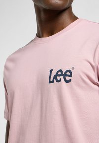 T-shirt di cotone rosa chiaro con logo "Lee" blu navy sul petto, con scollo rotondo classico e maniche corte.