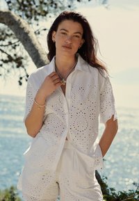 Completo in cotone bianco con una camicia a maniche corte con motivo a occhiello e colletto, abbinato a pantaloni corti, ambientato in uno sfondo costiero.