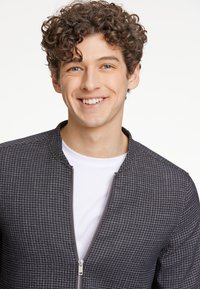 Grå rutigt bomberjacka med dragkedja framtill, i kombination med en vit T-shirt under. Tyget har en subtil textur och en slät finish.