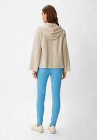 Maglione cappuccio beige con texture a coste e maniche ampie, abbinato a pantaloni slim-fit azzurri e sneakers bianche.