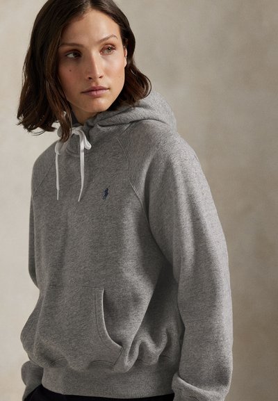 Frau mit schulterlangem braunem Haar, die einen grauen Kapuzenpullover mit weißen Kordeln und einem kleinen marineblauen Logo auf der Brust trägt.