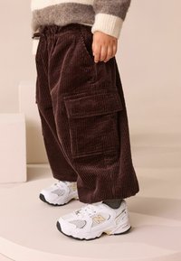 Bruna kordbyxor med sidofickor, i avslappnad passform, tillsammans med vita och guldiga löparskor. Texturerad tyg synlig.