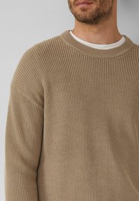 Beiger Strickpullover mit gerippter Textur und Rundhalsausschnitt; schulterfallen und lockere Passform, über ein weißes Hemd getragen.