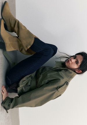 Žena v zelenej trenčkovej bunde a tmavých džínsoch sa opiera dozadu s nohami zdvihnutými na stene, obutá v pieskovo hnedej semišovej členkovej obuvi.