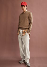 Brauner Strickpullover über einem karierten Orange- und Weißhemd, hellgraue Hose, graue Sneaker und eine rote Mütze mit grafischem Logo.