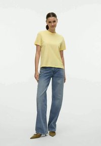 Geel T-shirt met korte mouwen, relaxed fit, gecombineerd met lichtblauwe flared jeans. Model draagt puntige schoenen en staat tegen een effen achtergrond.