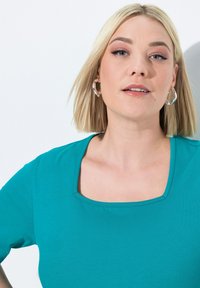 Tyrkysový top s krátkým rukávem a čtvercovým výstřihem. Vyroben z měkké tkaniny, má jemnou strukturu a čisté linie. Viditelné stříbrné kroužkové náušnice.