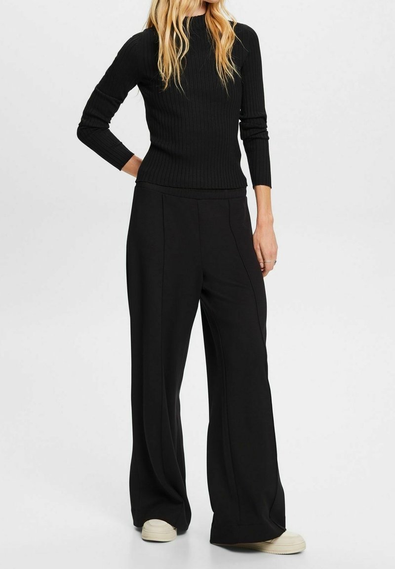 Femme portant un haut noir à manches longues côtelé et un pantalon noir à jambes larges avec des baskets blanches, debout devant un fond clair uni.