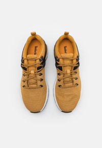 Žluté textilní tenisky s černými akcenty, kulatou špičkou a gumovou podrážkou. Disponují vzorovanou texturou a černými tkaničkami. Vnitřní branding Timberland.