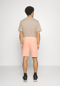 T-shirt beige à manches courtes et short rose avec une poche arrière, porté avec des baskets noires et des chaussettes sombres. Design simple et décontracté.