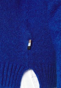 Modrý pletený svetr se žebrovaným lemem a malou nášivkou se třemi horizontálními barevnými pruhy: černým, bílým a hnědým.