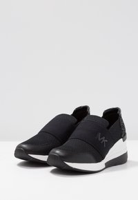 Sneakers neri senza lacci in tessuto a rete, con bande elastiche e un retro testurizzato. Presentano una suola intermedia bianca con un motivo geometrico e un dettaglio del logo.