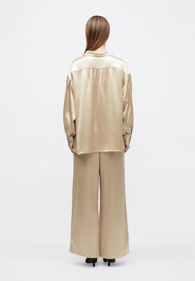 Žena stojaca chrbtom, oblečená v voľnej béžovej saténovej blúzke a zodpovedajúcich širokých nohaviciach s čiernymi špicatými podpätkami.