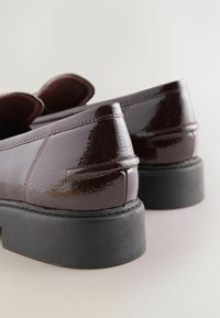 Burgundi värvi patendinahast loafereid läikiva viimistlusega, millel on lame must kummitald ja peene tikandiga detail ülaosas.