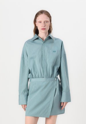 Nuori nainen, joka käyttää vaaleansinistä, pystyraitakuosista paitamekkoa, jossa on pitkät hihat, suuret kalvosimet ja kietaisuhameen etuosan yksityiskohta.