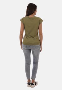 Donna con lunghi capelli castani che indossa una maglietta a maniche corte verde oliva, jeans grigio chiaro e scarpe da ginnastica bianche, vista di spalle.