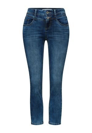 Persoon draagt een blauwe jeans met hoge taille en dubbele knoopsluiting, wit overhemd in de broek gestopt en een roze jas met gouden knopen.