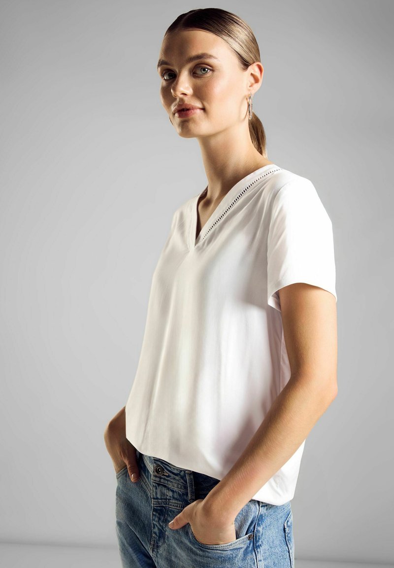 Blusa bianca a maniche corte con scollatura a V e texture leggera, caratterizzata da un design semplice lungo il colletto. Abbinata a jeans in denim blu.