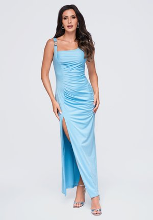 Femme aux longs cheveux foncés portant une robe bleue claire sans manches avec fronces, une fente à la cuisse et des sandales à talons hauts argentées, debout sur un fond blanc.