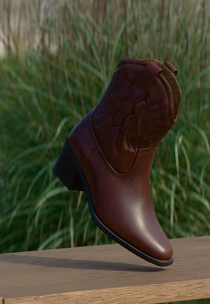 Stivale da cowboy in pelle marrone con cuciture decorative, inclinato verso l'alto su una superficie di legno con sfondo sfocato di erba verde.