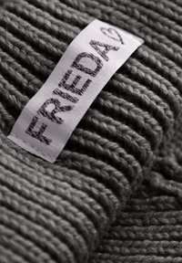Grijze gebreide stof met een geribbelde textuur en een wit label met de tekst "FRIEDA" in donkere draad gestikt.