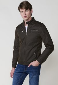 Rjav zip-up jopič z dvignjenim ovratnikom, kvačkanimi ramenskimi poudarki in dvema žepoma s zadrgo na prsih. Obdan z belo majico in denim jeans hlačami.