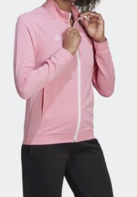 Chaqueta deportiva de color rosa claro con cremallera, cuello alto y mangas largas. Presenta bolsillos laterales y un sutil detalle de logo en el pecho.