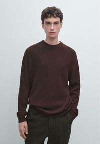 Brązowy sweter dzianinowy z okrągłym dekoltem, długimi rękawami, ściągaczami na mankietach oraz miękką fakturą, zestawiony z ciemnymi spodniami w paski.