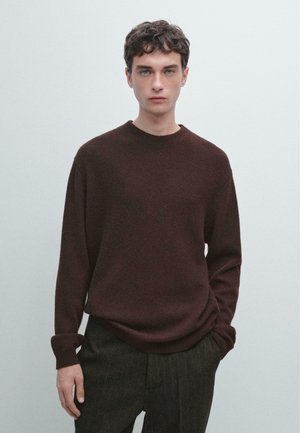 Maglione marrone a maglia con scollo rotondo, maniche lunghe, polsini a costine e una consistenza morbida, abbinato a pantaloni scuri a righe.