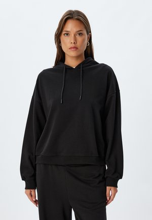 Haut à capuche noir en tissu doux, avec une poche kangourou sur le devant, une capuche avec cordon de serrage et des poignets côtelés. Associé à un pantalon noir assorti.