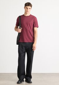 Jonge man die staat te poseren in een bordeauxrode Hugo t-shirt, zwarte wijde broek, zwarte schoenen, en een zwarte schoudertas in de hand.