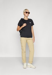 Črn majica s kratkimi rokavi in logotipom, khaki cargo hlače in svetli športni copati. Dodatki: rjave sončne očala, črn nahrbtnik z naramnicami.