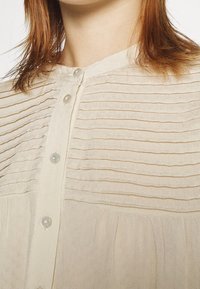 Béžová texturovaná blůza s horizontálními sklady na horní části hrudníku, zapínání na knoflíky s malými kulatými průhlednými knoflíky, nošena osobou.