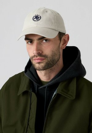 Mand med kort skæg iført beige baseballkasket, sort hættetrøje og olivengrøn jakke mod ensfarvet baggrund.