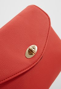 Clutch bag in pelle rossa testurizzata con pattina arrotondata e chiusura ovale dorata. Presenta cuciture visibili e un rivestimento interno liscio.