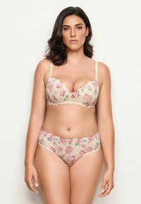 Žena s polodlouhými tmavými vlasy, oblečená v ladícím květinovém krajkovém podprsenku a kalhotkách, stojící na jednoduchém světlém pozadí.