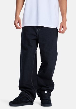 Homme portant un jean noir ample, des baskets noires DC et une chemise blanche, debout devant un fond clair uni.