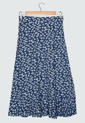 Mujer con blusa blanca de manga corta, falda midi azul con estampado floral blanco y sandalias blancas de tacón, de pie sobre un suelo de madera.