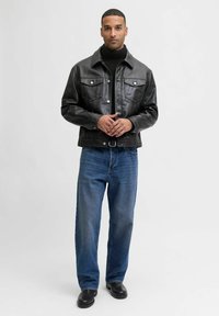 Man in een zwarte leren jas, zwarte coltrui, blauwe spijkerbroek en zwarte schoenen, staand tegen een effen witte achtergrond.