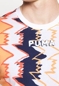 Krátký rukáv tričko v bílé barvě s oranžovými a námořnickými zigzagovými vzory. Na přední straně je napsáno logo "PUMA" silnými bílými písmeny. Materiál: bavlna.