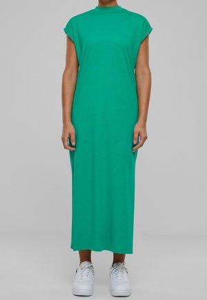 Personne portant une robe verte sans manches longueur cheville associée à des baskets blanches, debout devant un fond gris uni.