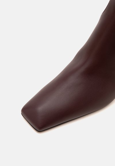 Gros plan d’une botte en cuir marron foncé avec un design à bout carré sur un fond clair uni.